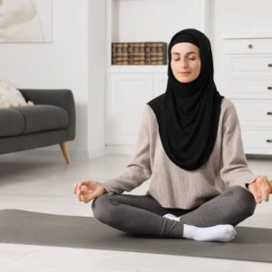 Muslim woman in hijab meditating on fitness mat at home. Space for text